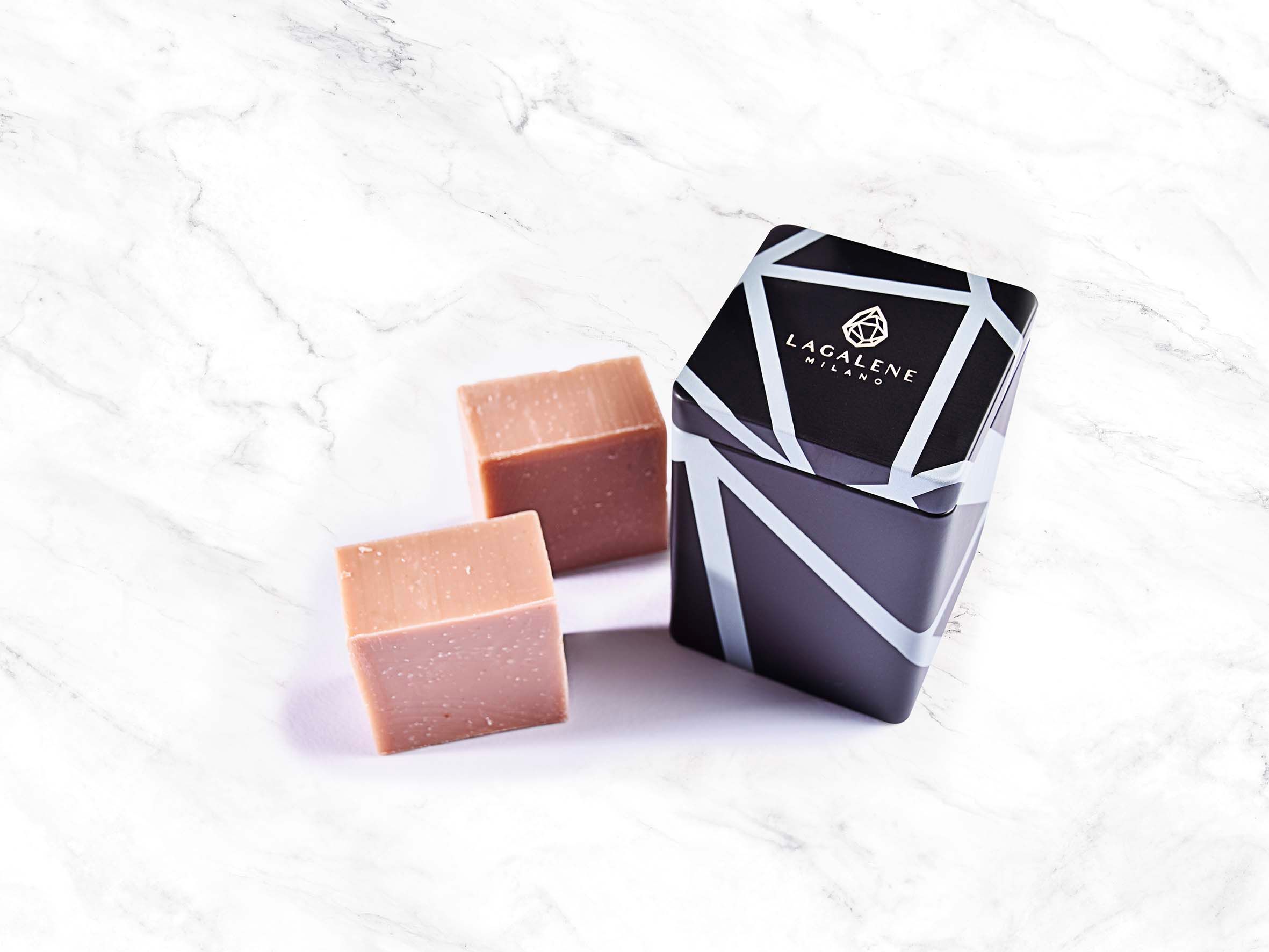 Deux savons à côté d'une boîte à motifs noirs et gris. La boîte affiche le nom de la marque Lagalene Milano.