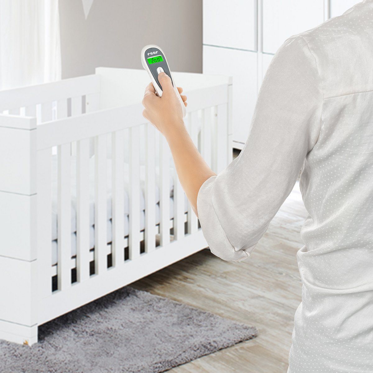 Personne tenant un thermomètre infrarouge sans contact. Marque : reer. Appareil pointé vers un lit bébé.