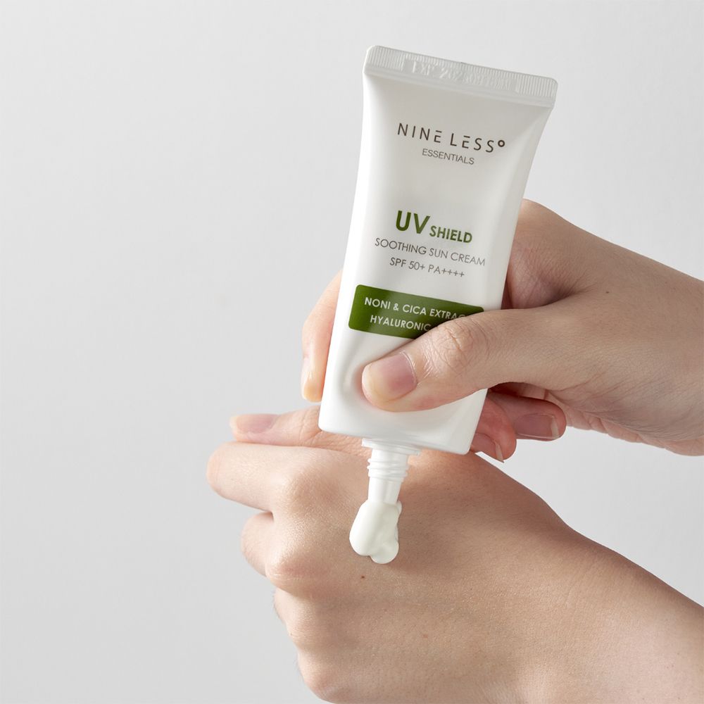 Witte tube wordt op een hand gedrukt. Opschrift: UV Shield, SPF 50+ PA++++, Noni & Cica Extract, Hyaluronzuur. Merk: NINE LESS.