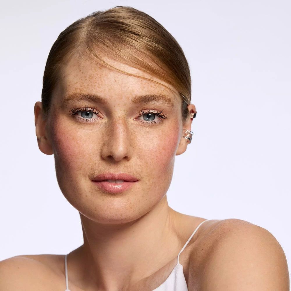 Portrait d'une femme avec du blush rose sur les joues. Elle porte un haut blanc. Des taches de rousseur sont visibles.