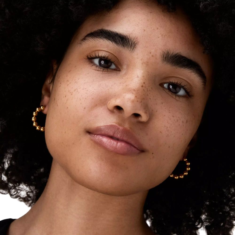 Portrait d'une femme avec des taches de rousseur. Elle porte des boucles d'oreilles dorées. Regard vers la caméra.