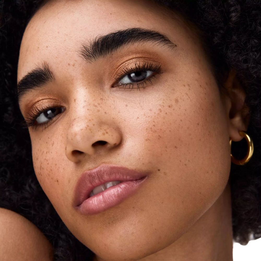 Close-up van een gezicht met gouden oogschaduw. De persoon heeft sproeten en draagt gouden oorringen.
