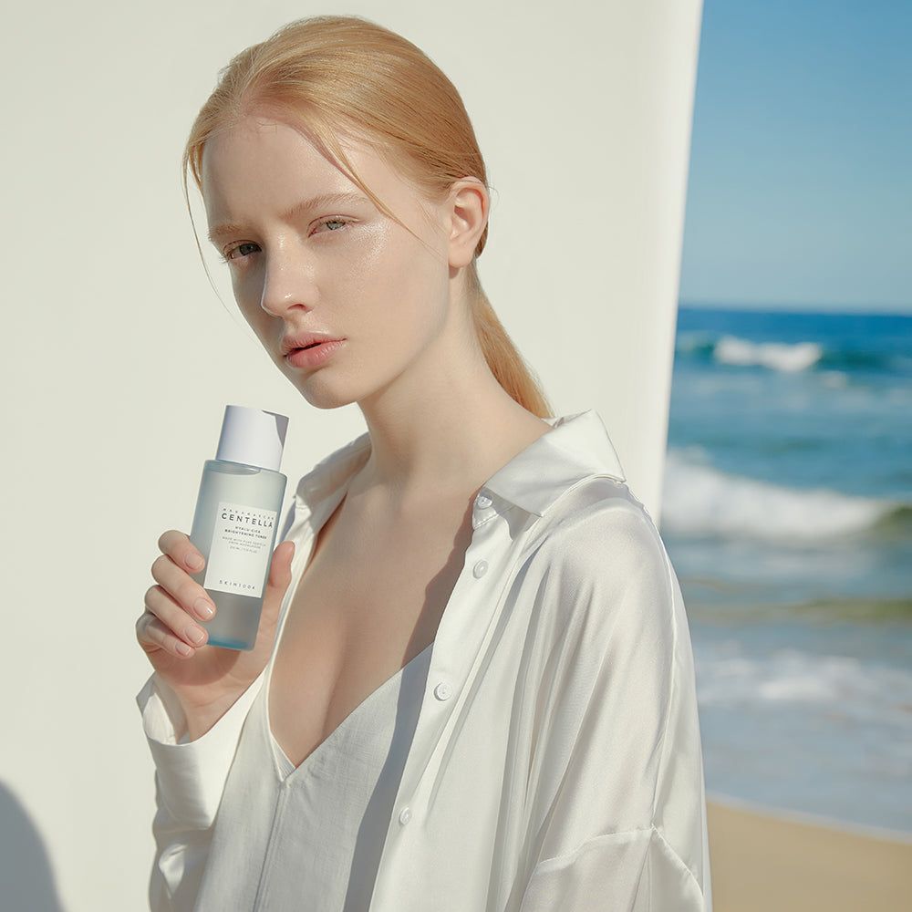 Vrouw houdt een fles vast. Opschrift: MADAGASCAR CENTELLA, HYALU-CICA BRIGHTENING TONER. Achtergrond: strand en zee.