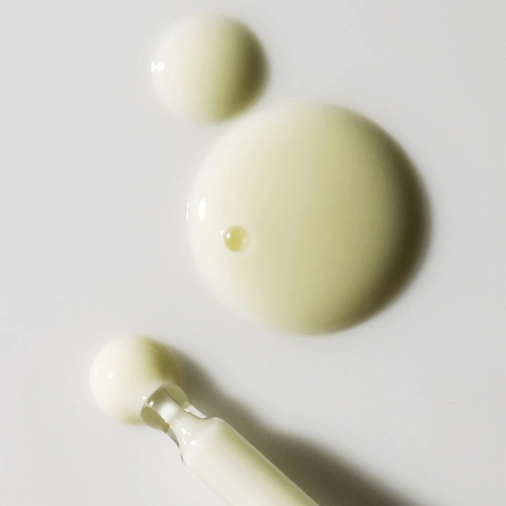 Trois gouttes de sérum, dont une avec une pipette. Liquide jaune clair. Pipette avec manche blanc.