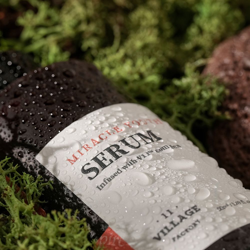 Bruine serumfles met wit etiket, bedekt met waterdruppels. Tekst: MIRACLE YOUTH SERUM. VILLAGE 11 FACTORY. Op mos.