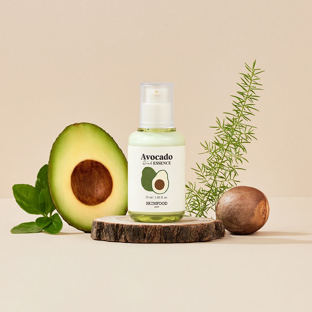 Serumfles op houten schijf, naast avocadohelft, bladeren en zaden. Opschrift: Avocado Rich Essence, SKINFOOD.