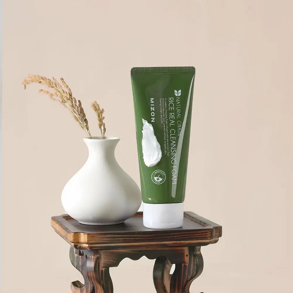 Groene tube met witte dop en schuim. Opschrift: MIZON, Rice Real Cleansing Foam. Logo en tekst: Natural Grain. Op een tafel.