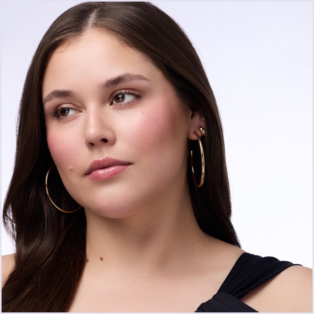 Portrait d'une femme avec une peau bronzée et du blush rose sur les joues. Elle porte des créoles dorées.