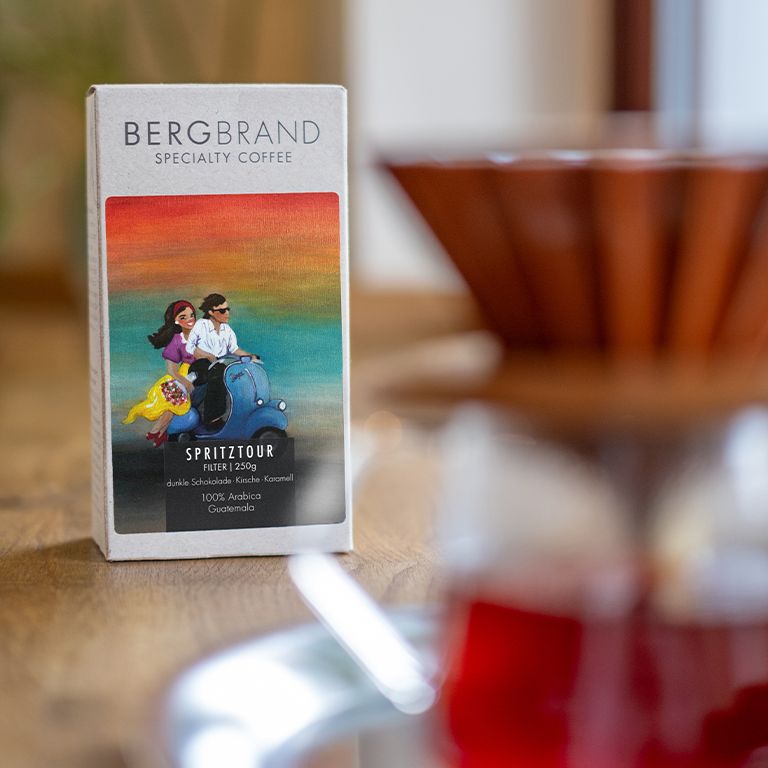 Paquet de café BERGBRAND Spritztour à côté d'une cafetière. Illustration de personnes sur un scooter.