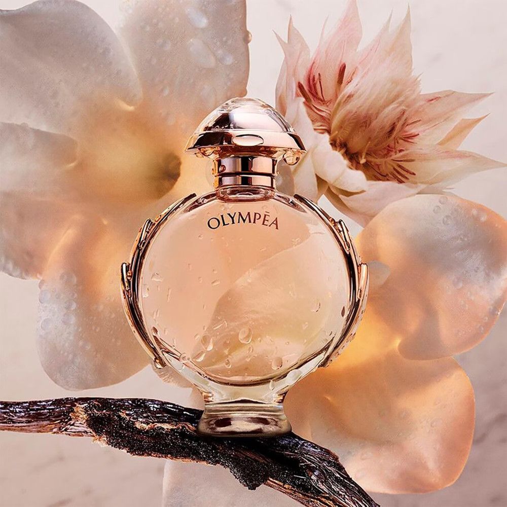 Parfumfles, rond, roze-goudkleurig. Geplaatst op een tak, omringd door bloemen en bladeren. Druppels op het glas.