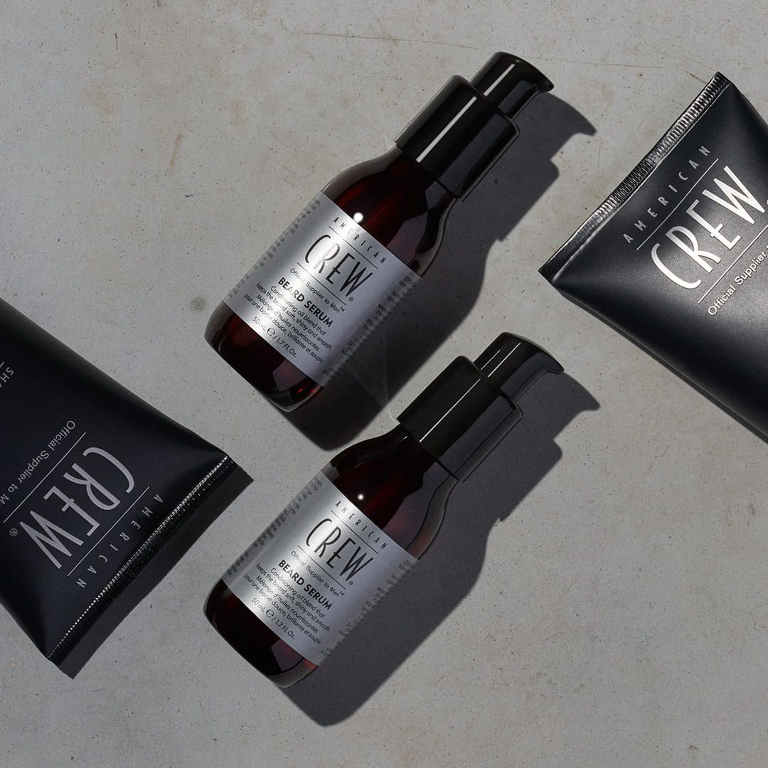 Deux flacons et deux tubes de sérum à barbe AMERICAN CREW sur fond gris. Emballage noir avec texte argenté.