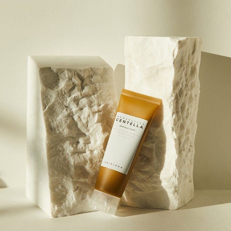 Beige tube met wit etiket. Opschrift: MADAGASCAR CENTELLA AMPULE FOAM. Merk: SKIN1004. Naast twee witte blokken.