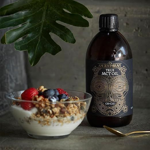 Un bol de yaourt, muesli et baies est placé à côté d'une bouteille d'huile MCT. Une petite cuillère dorée est à côté.