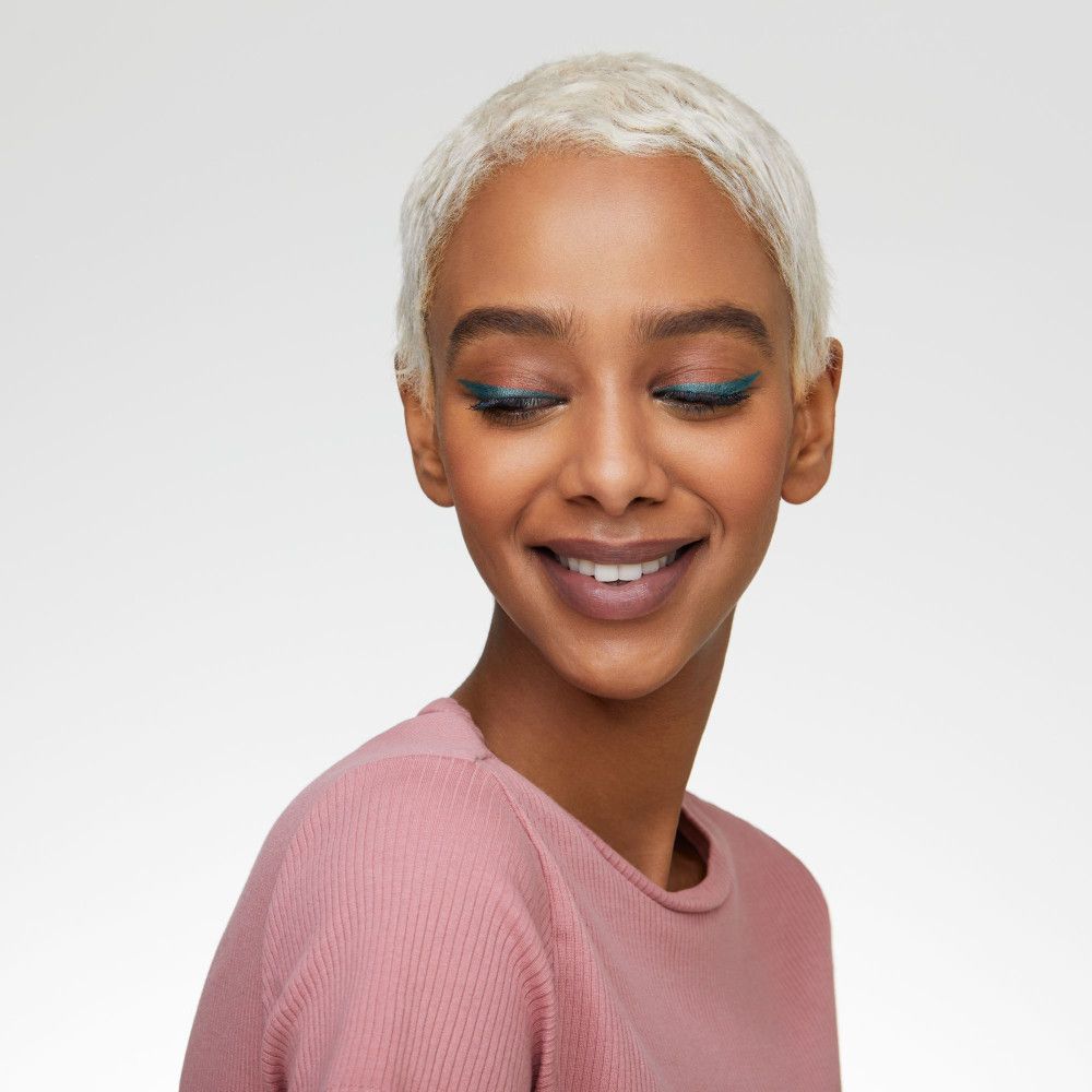 Femme aux cheveux blonds avec trait de kajal. Elle porte un haut rose et sourit.