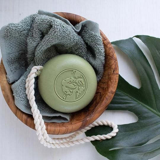 Ronde groene zeep met koord in een houten kom, naast een handdoek en een blad.