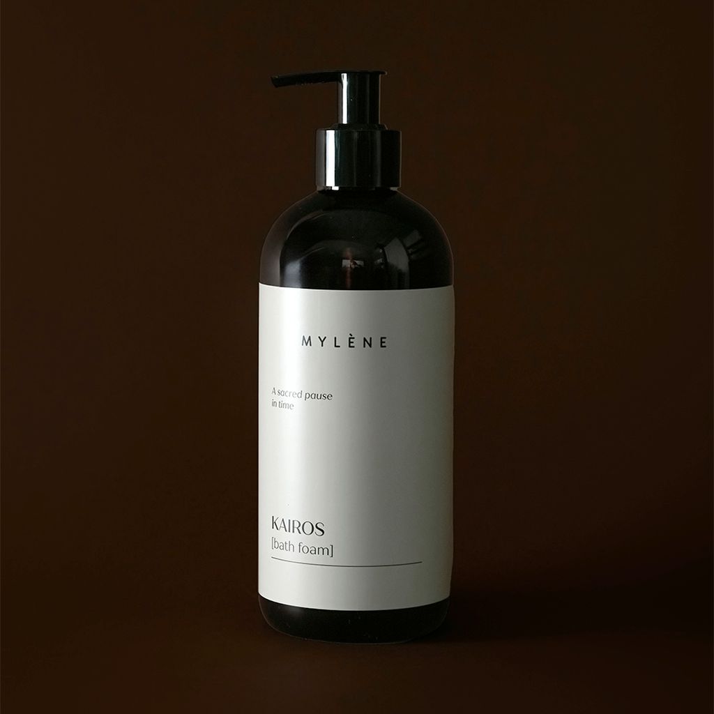 Donkere pompfles met licht etiket met "KAIROS [Bath foam]" erop, tegen een bruine achtergrond.