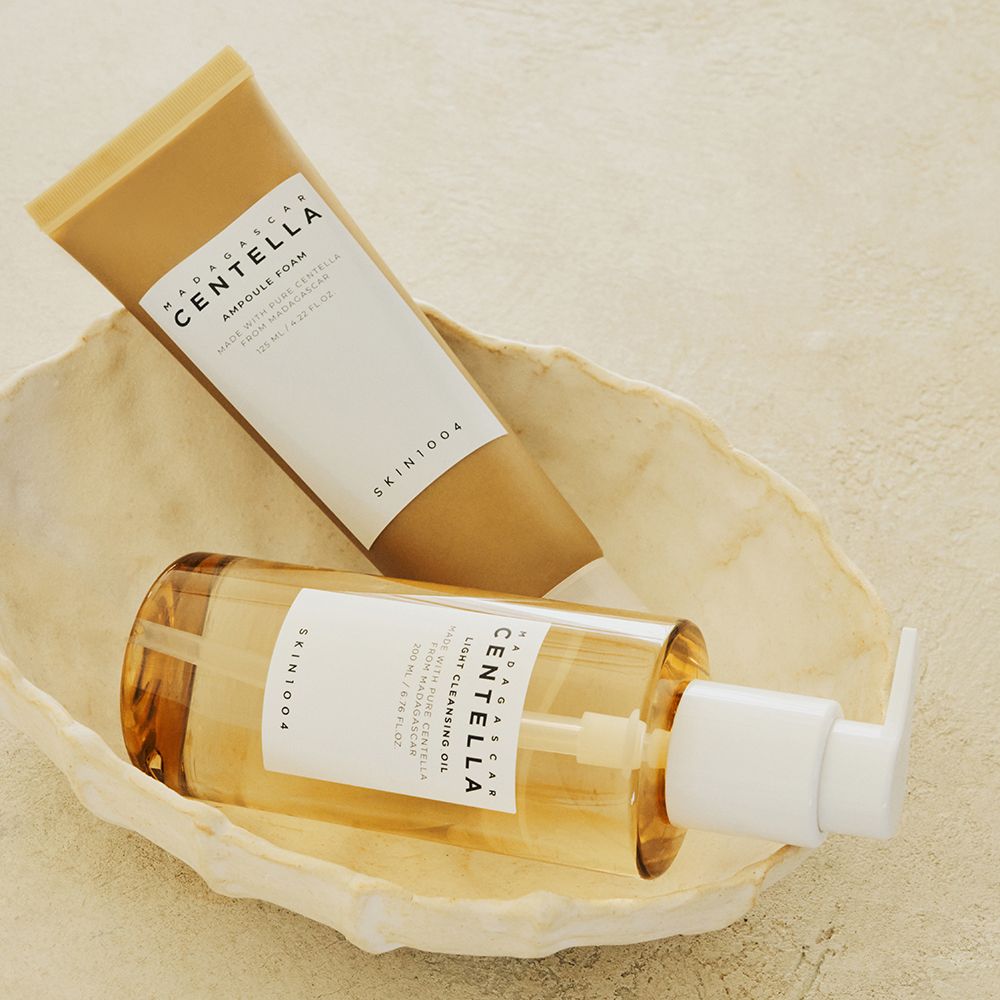 Deux produits dans un bol crème. Une bouteille d'huile nettoyante avec pompe et un tube de mousse ampoule. Les deux produits portent la marque SKIN1004.