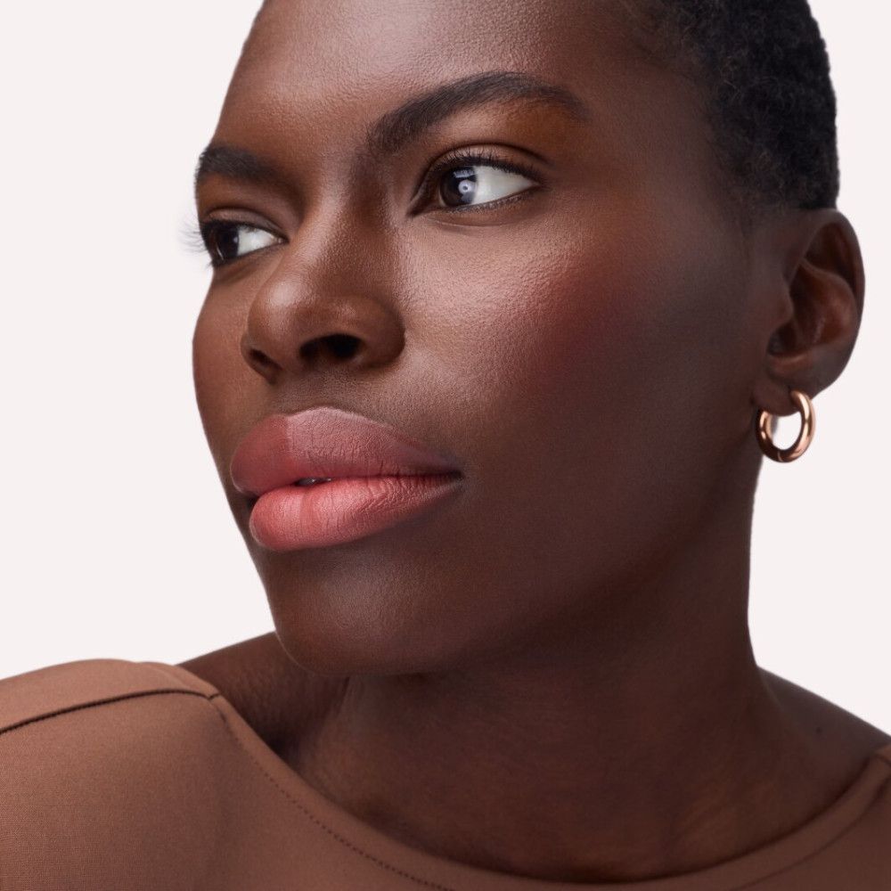 Portrait d'une femme avec des lèvres roses. Elle porte des créoles dorées et un pull marron.