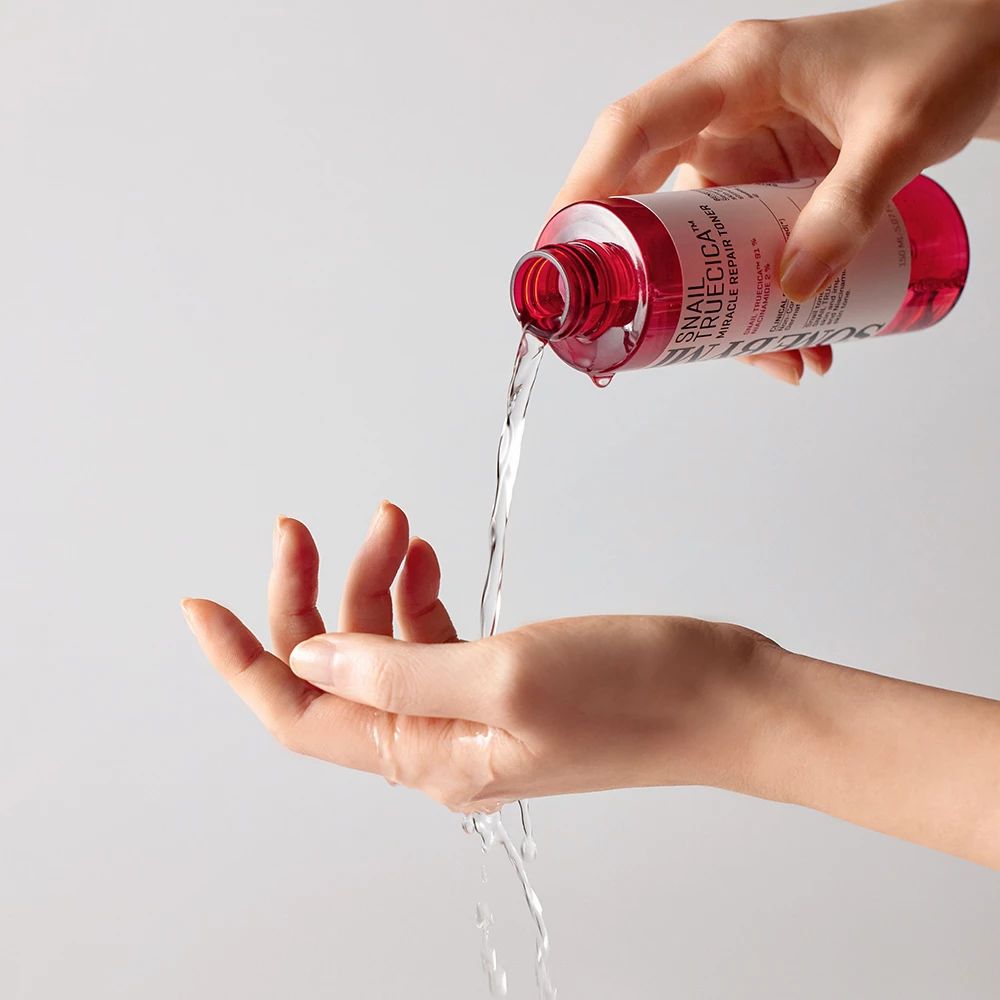 Une main verse du liquide d'une bouteille rouge sur une autre main. La bouteille porte l'inscription SOME BY MI.