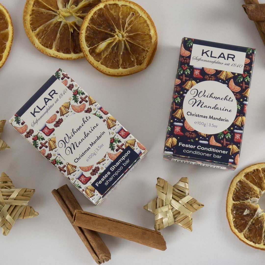 Deux emballages Klar-Seifen avec décor de mandarine et de Noël. Bâtons de cannelle, tranches d'orange séchées et étoiles de paille à côté.