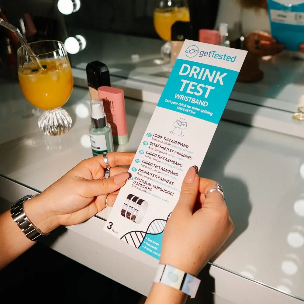 Persoon houdt een verpakking vast met "GetTested Drink Test Wristband". Meerdere armbanden zijn zichtbaar. Achtergrond: kaptafel.