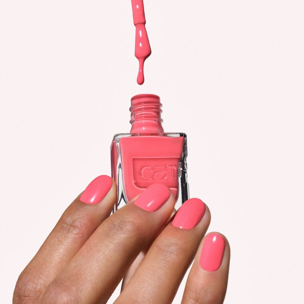 Een hand met koraalrode nagellak houdt een fles nagellak vast. Een kwastje laat kleur druppelen.