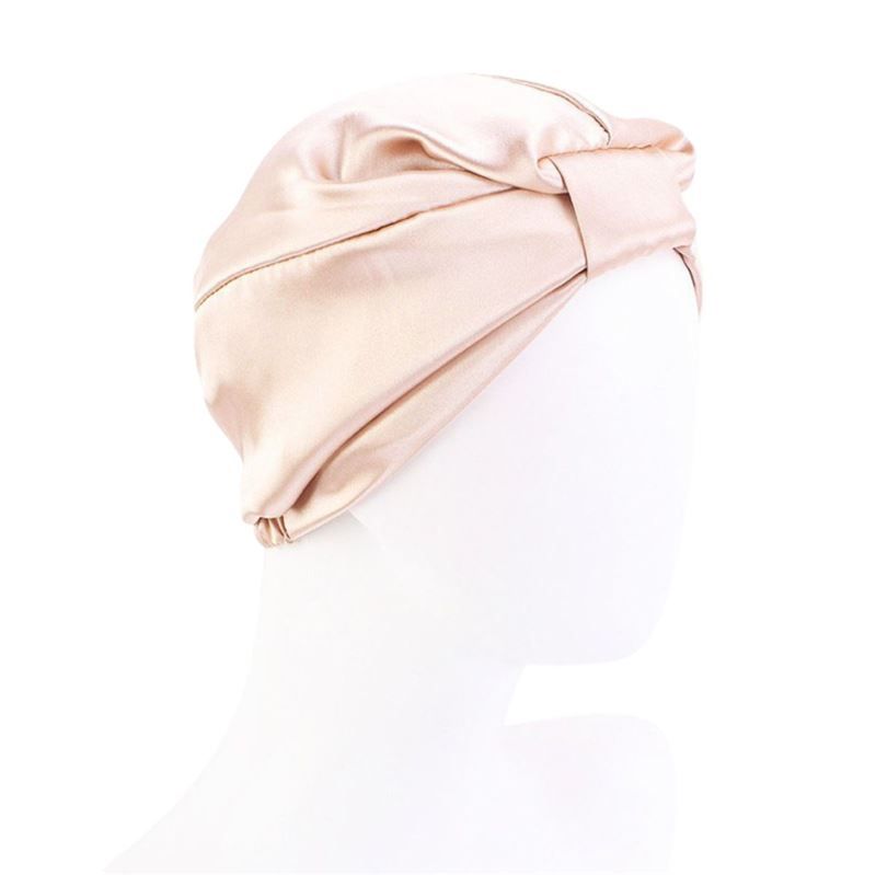 Un bonnet de nuit en soie de couleur rose pâle. Le bonnet a une forme de turban avec un nœud décoratif à l'avant. Le matériau semble lisse et brillant.