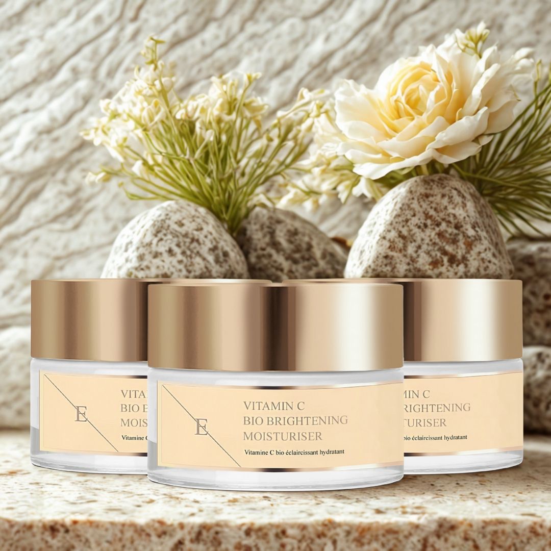 Trois pots avec couvercles dorés, placés sur des pierres, avec des fleurs. Les étiquettes indiquent : Vitamin C Bio Brightening Moisturiser.