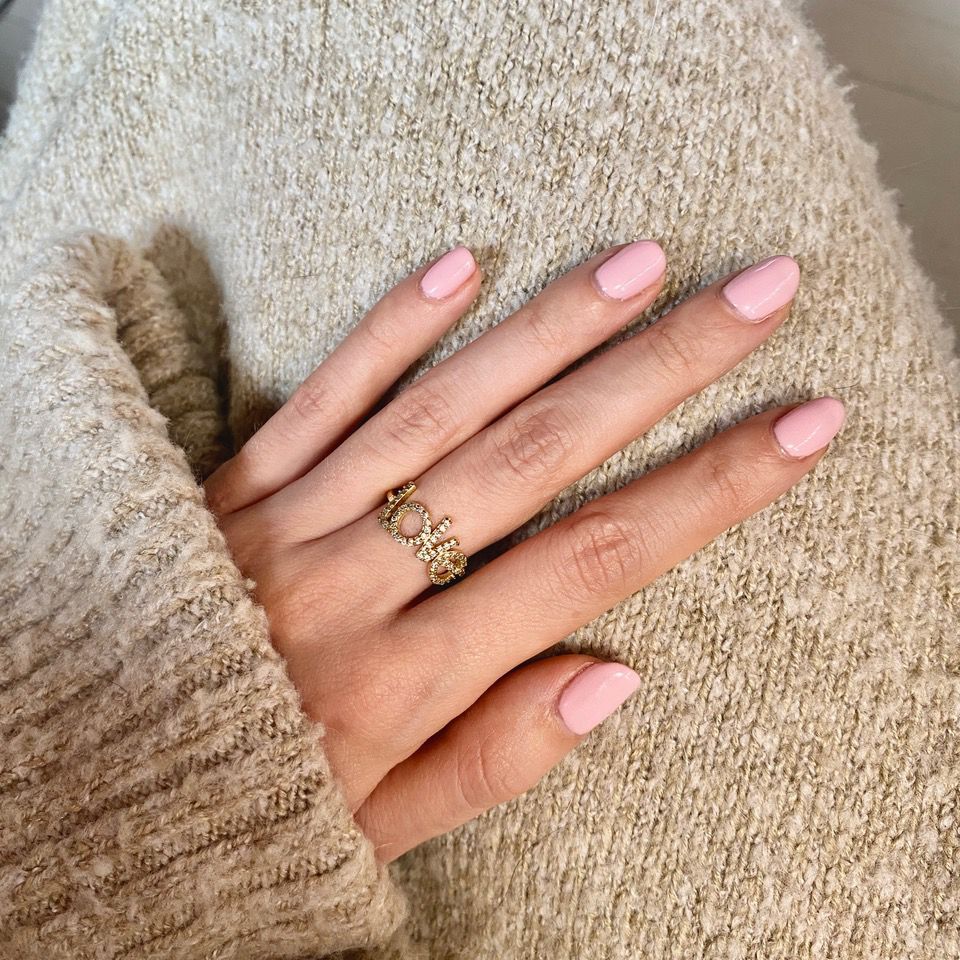 Main avec ongles roses ovales. La main porte une bague dorée. La main porte un pull beige.