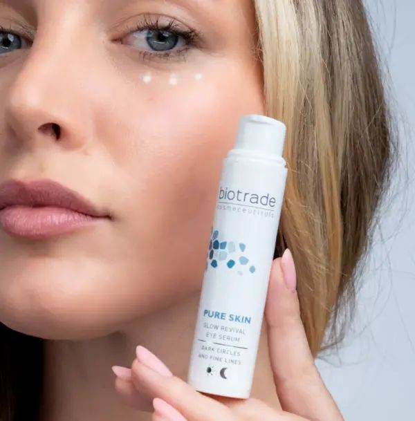 Femme tenant un flacon de crème pour les yeux Biotrade Pure Skin. Flacon blanc avec motif bleu. Texte : Pure Skin Glow Revival Eye Serum.
