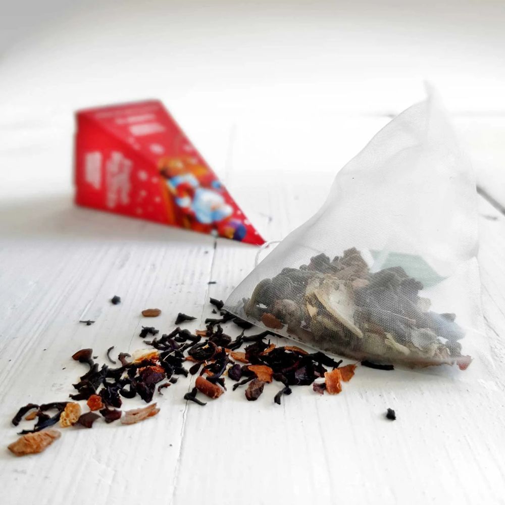 Theezakje met losse blaadjes. Daarnaast een driehoekige verpakking. Achtergrond: houten plank met onscherp rood cadeau.