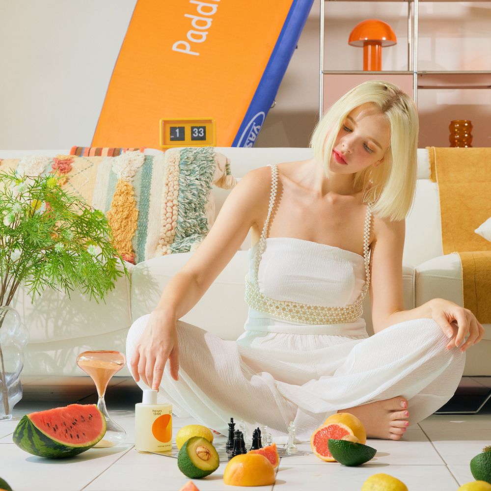 Een vrouw zit op de grond met fruit en een fles douchegel. Tekst en een oranje ontwerp zijn zichtbaar op de fles.