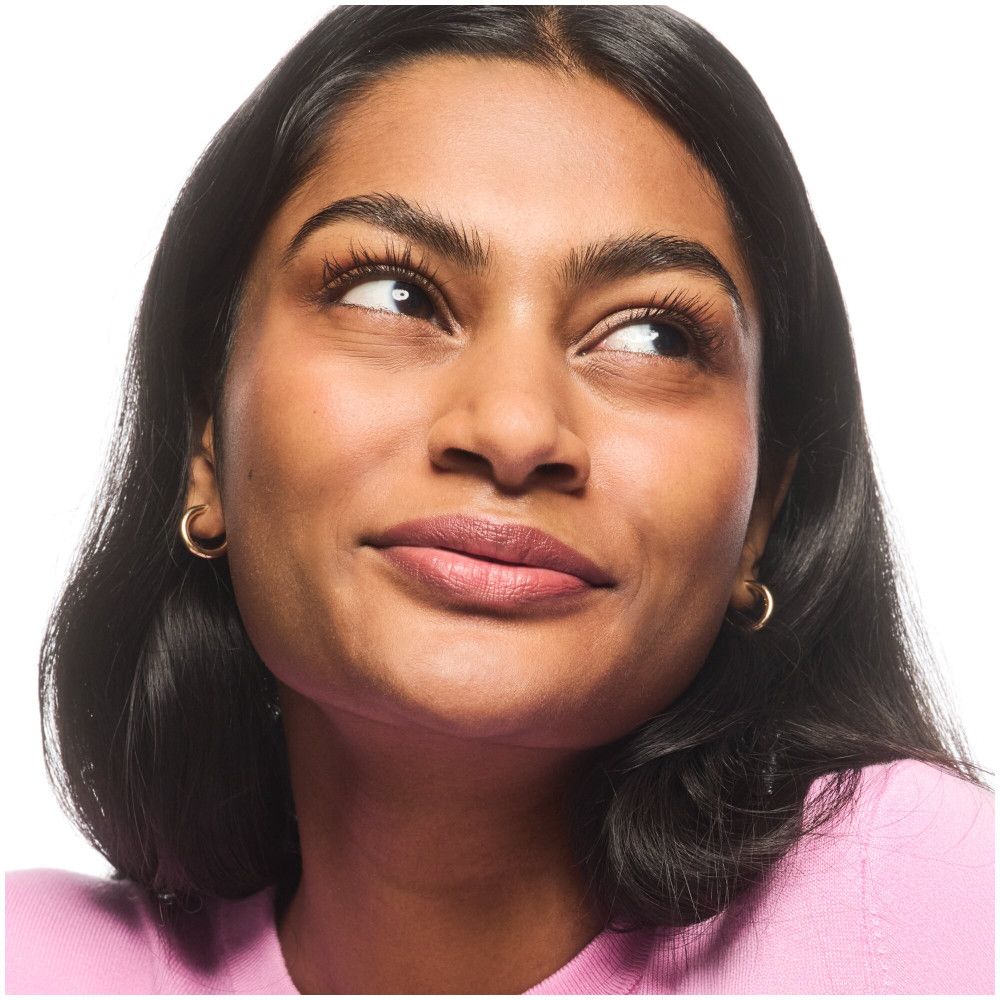 Portrait d'une femme aux cheveux foncés regardant vers le haut. Elle porte un pull rose et des boucles d'oreilles dorées.