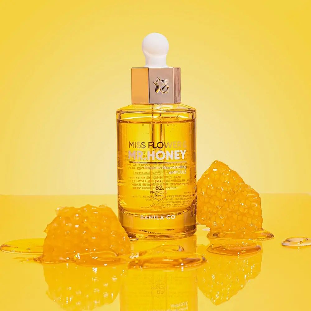 Glazen fles met goudkleurig serum. Opschrift: Miss Flower & Mr. Honey. Honingraten ernaast. Gele achtergrond.