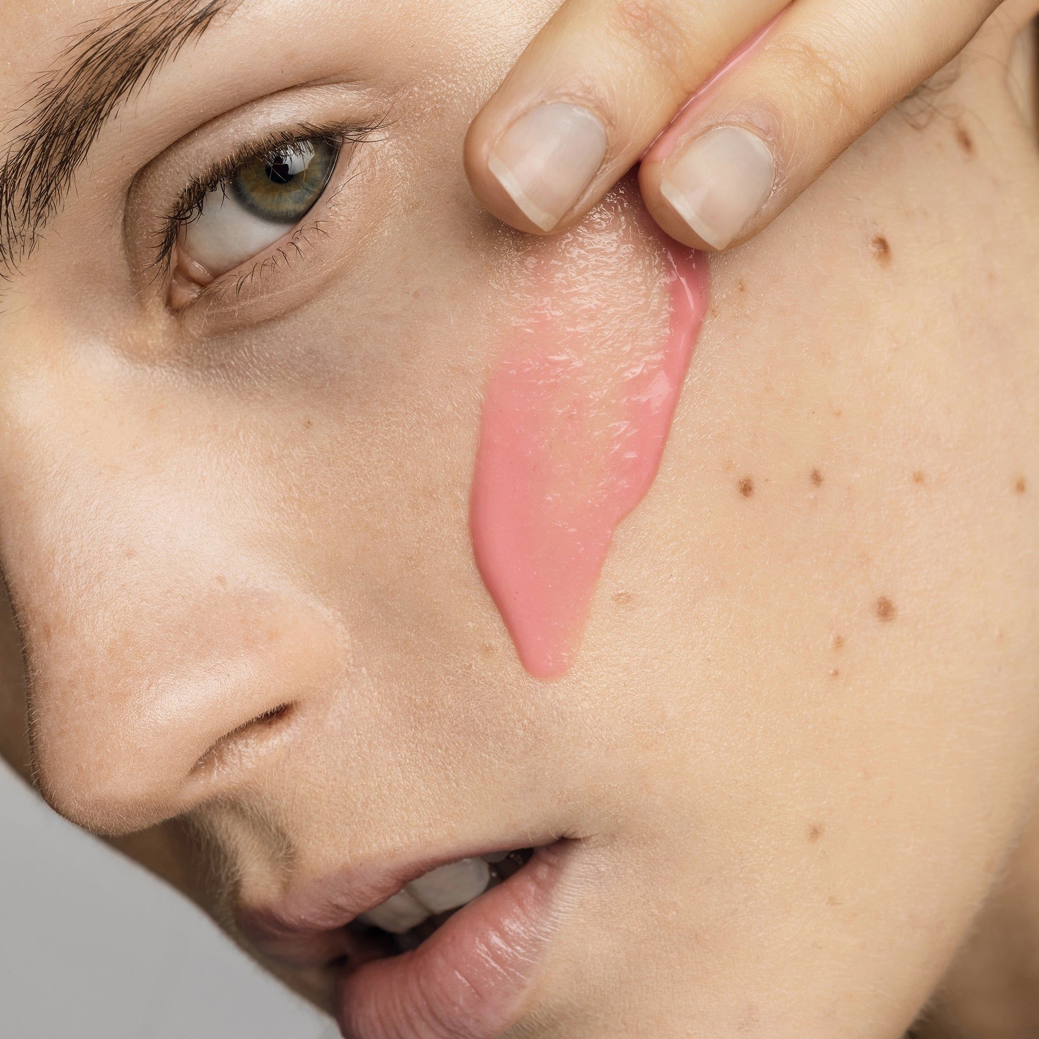 Close-up van een gezicht. Roze serum wordt op de wang aangebracht. Huid met sproeten.