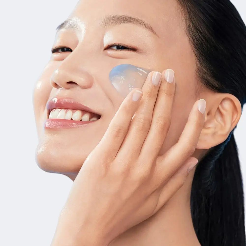 Femme appliquant une crème bleue sur son visage. Elle sourit. Peau claire, cheveux foncés.