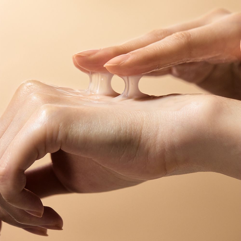 Mains avec produit. Le produit est transparent et gélatineux. Fond : beige.