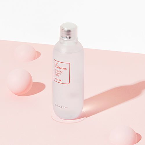 Fles met rood etiket: AC Collection, Calming Liquid Mild, COSRX. Op roze oppervlak. Drie roze ballen.