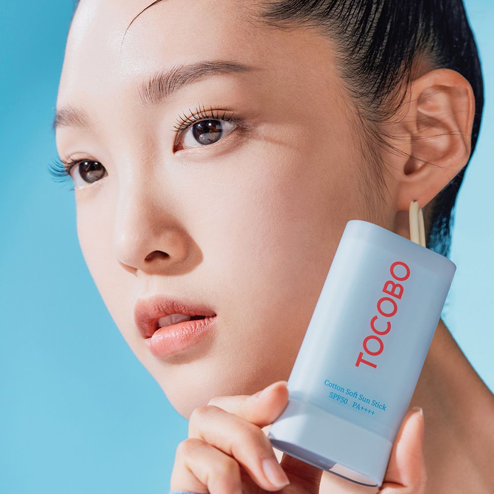 Portret van een vrouw die een zonnebrandstick vasthoudt. Rode tekst TOCOBO. Tekst: Cotton Soft Sun Stick SPF50+ PA++++.