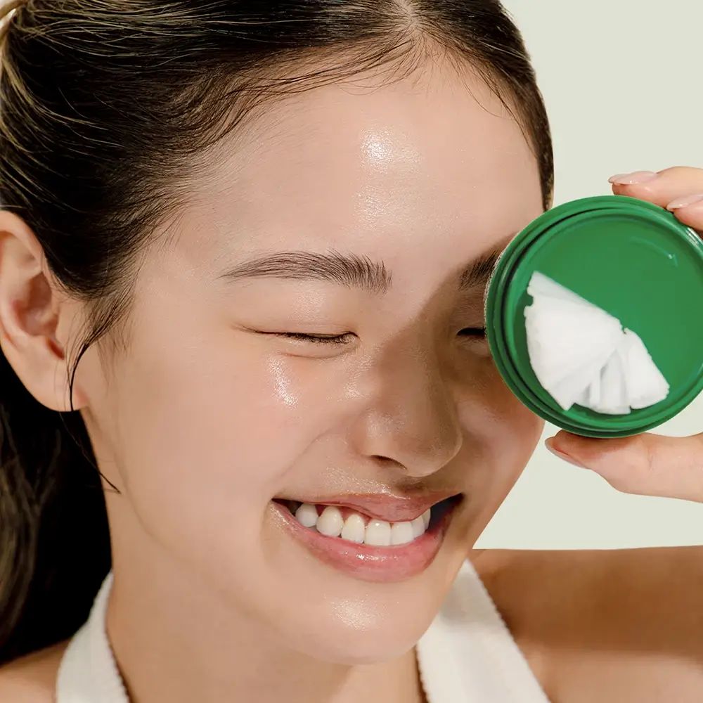 Femme tenant un couvercle vert avec baume nettoyant blanc. Visage à la peau brillante. Yeux fermés, souriant.