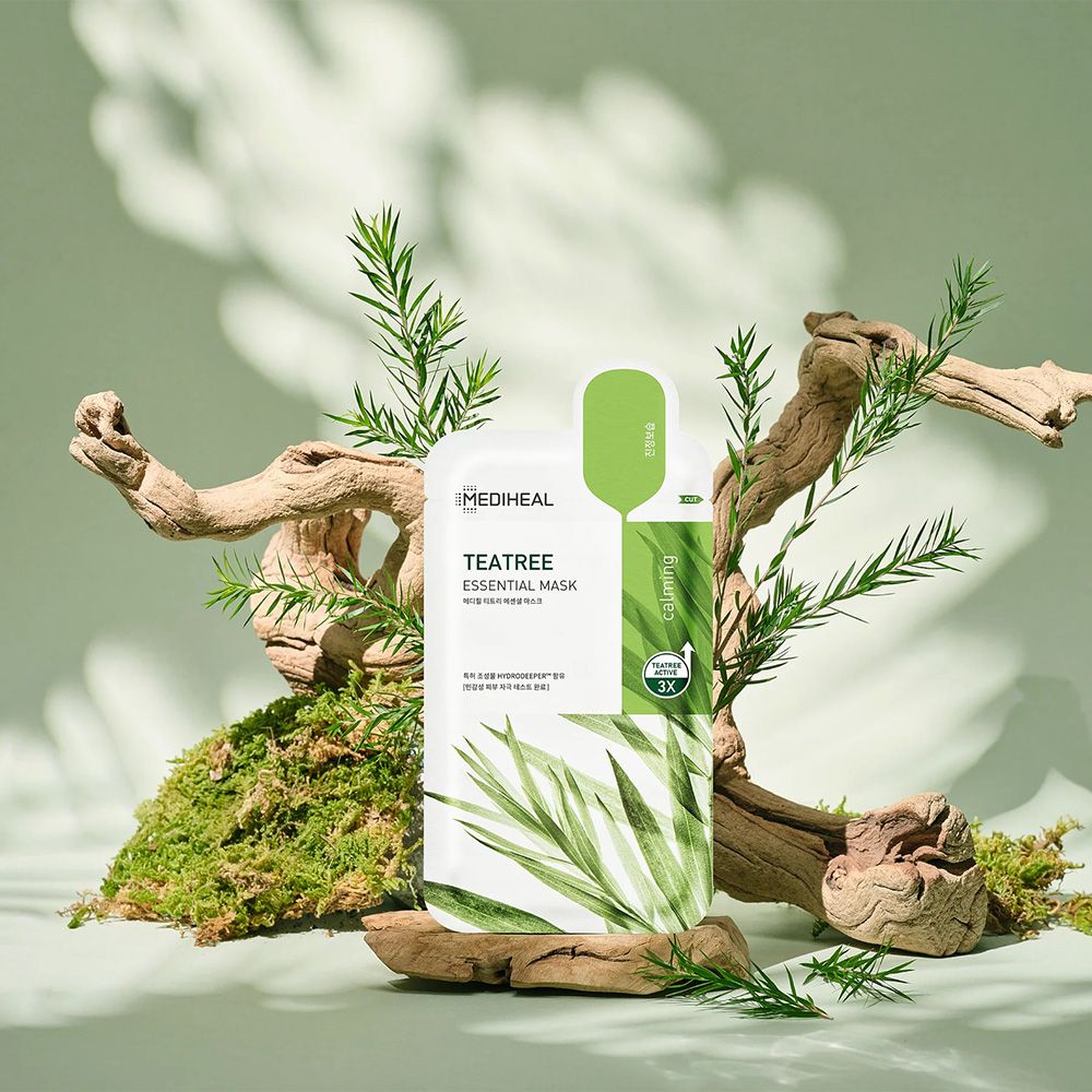 Emballage avec masque, entouré de bois et de plantes. Inscription: Tea Tree Essential Mask. Bande verte avec texte.