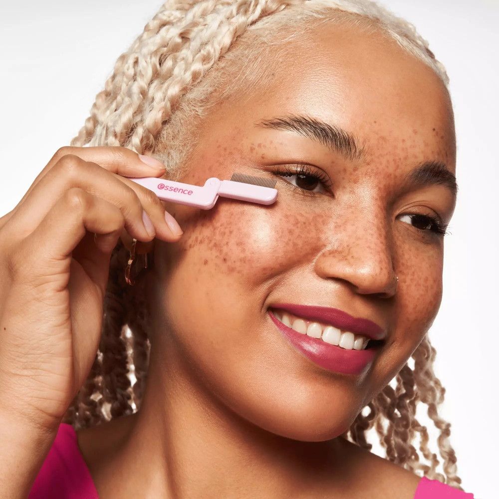 Femme aux boucles blondes utilisant un peigne à cils rose. Marque : Essence. Le peigne est placé sur l'œil.