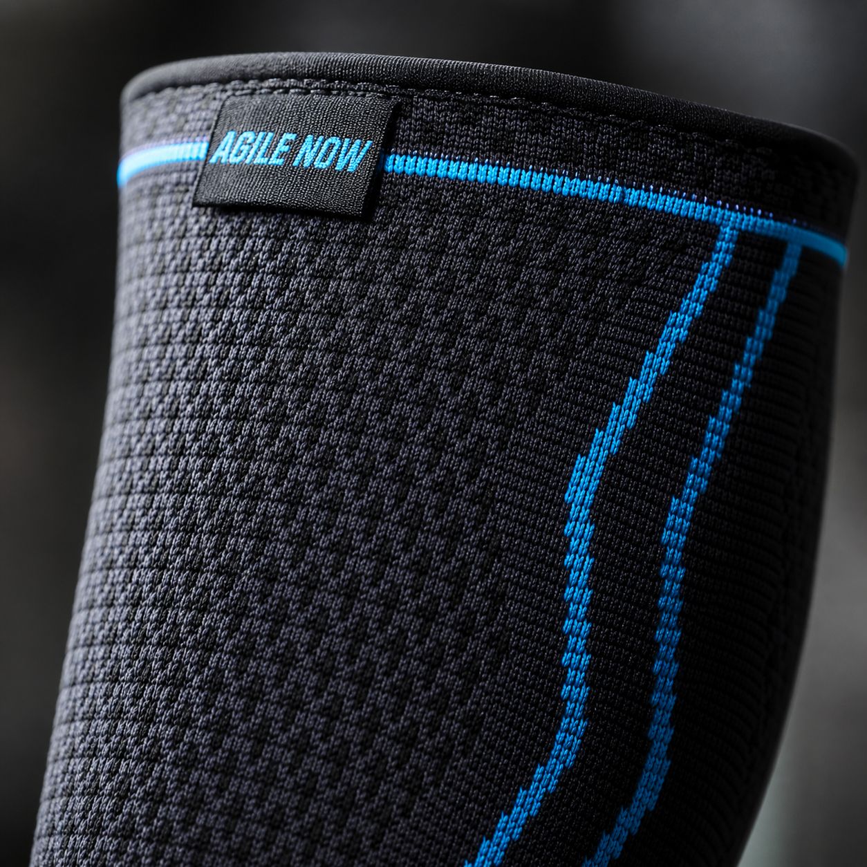 Close-up van een zwarte kuitbandage met blauwe lijnen en de tekst AGILE NOW. Zwarte achtergrond.