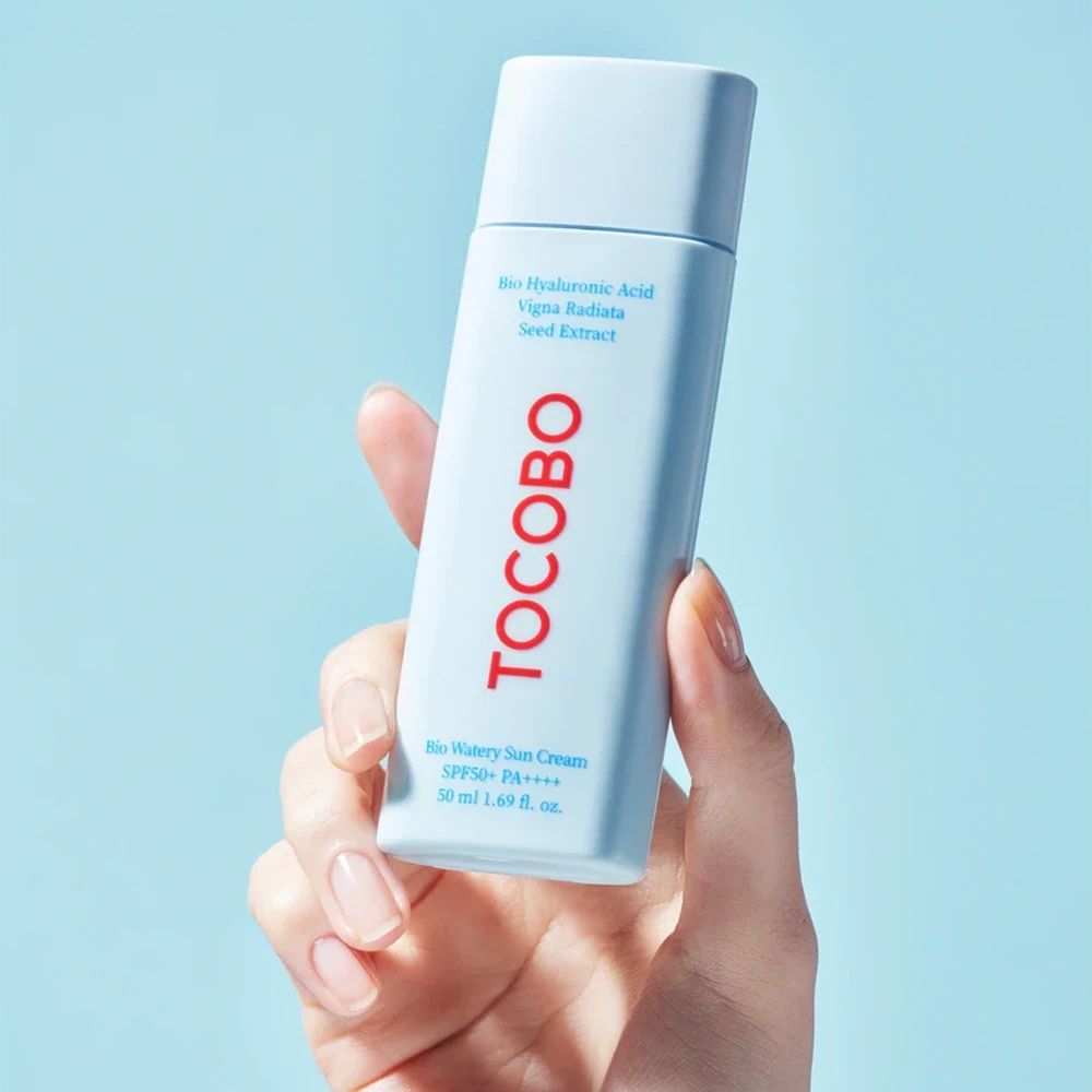 Main tenant un tube bleu clair. Mot TOCOBO en rouge. Texte : Bio Watery Sun Cream SPF50+ PA++++. 50 ml.