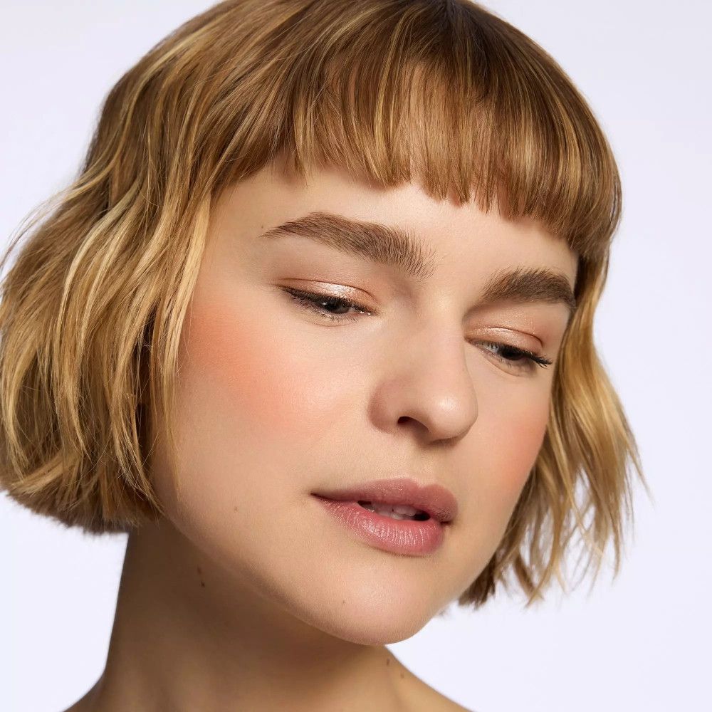 Femme aux cheveux blonds clairs et blush sur les joues. Elle regarde vers le bas.