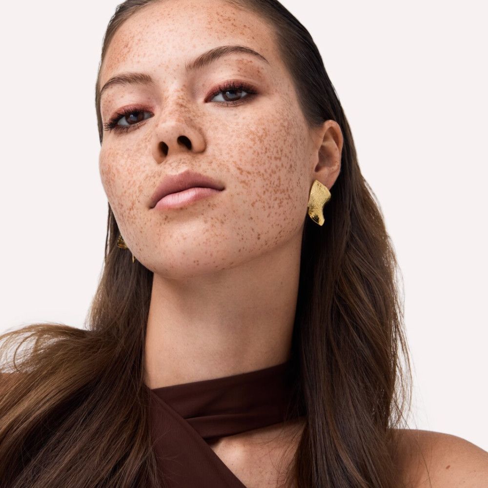 Portrait d'une femme avec des taches de rousseur. Elle porte des boucles d'oreilles dorées et du maquillage rouge pour les yeux.