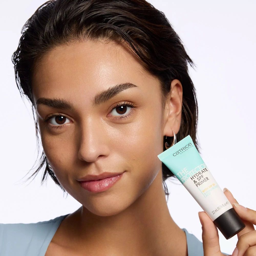 Femme tenant un tube. Inscription: Catrice, THE PROTECTOR HYDRATE & SPF PRIMER. 22 ml. Femme avec boucle d'oreille.