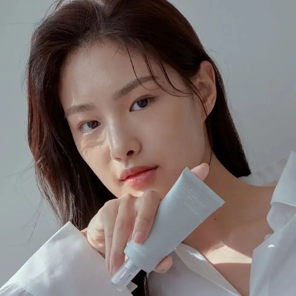 Femme tenant un tube gris. Texte illisible. Elle porte une chemise blanche. Elle regarde la caméra.