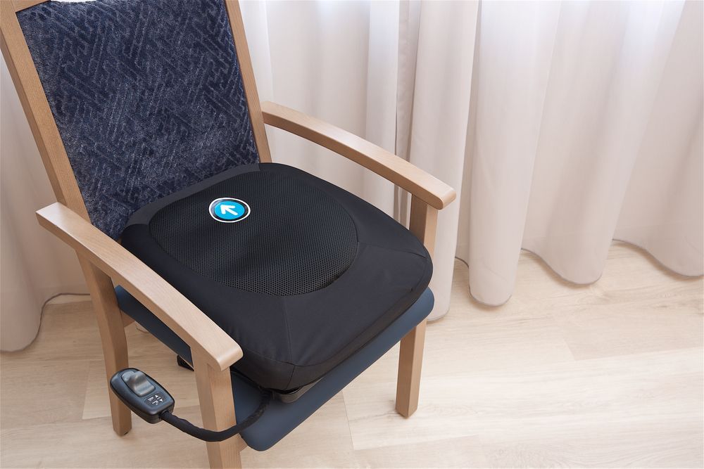 Zwarte elektrische opstaphulp op een stoel. Blauw logo zichtbaar. Afstandsbediening.