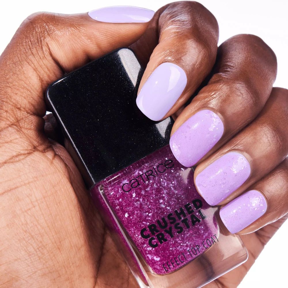 Main avec ongles vernis et flacon de vernis. Ongles lilas clair, vernis rose avec effet cristal concassé. Nom de la marque Catrice visible.